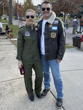 A woman and a man are dressed up as characters from the original "Top Gun" movie for Halloween. The woman wears aviator glasses and a pilot jumpsuit; the man, a pilot's jacket, aviator glasses, and blue jeans.