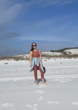 This photo shows a woman standing on a beach with blue sky overhead. She has long brown hair and wears sunglasses and a brown shirt and pants, with a blue or gray piece of clothing tied around her waist. She has a bag in one hand and is barefoot.