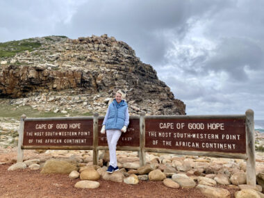 A woman in a light blue vest stands against a sign marking the Cape of Good Hope in South Africa.