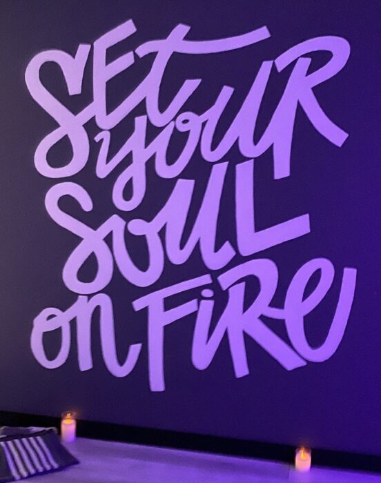 Light purple writing on a dark purple wall in a yoga studio reads "Set your soul on fire." Two candles on the floor are lit. 