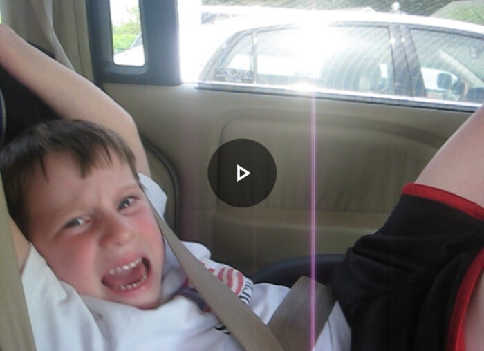 A screenshot of a video of a young boy in a car looking at the camera and screaming in agony, with his arms raised. He's squeezing down in the seat to avoid the sunlight shining through the car window. 