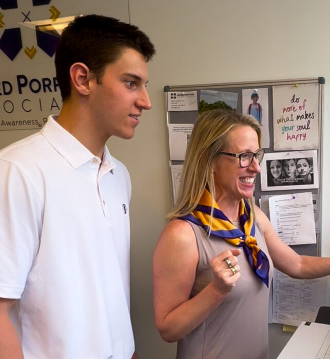 A side shot of a tall 19-year-old and his shorter mother as they look to the right of the frame and down and something while in an office. The mother is smiling excitedly.