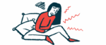 A person seated against a couple pillows is seen hunched with both arms clutching the stomach, in pain.