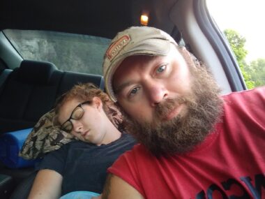 A man, sitting in the driver's seat of a car, takes a selfie of him and his wife, who is asleep in the back seat. He has a thick brown beard and is wearing a khaki hat and a red shirt. The woman has on dark-framed glasses and a navy shirt, and her head is resting on a pillow. We can't see much outside of the car, but there appear to be trees in the background.