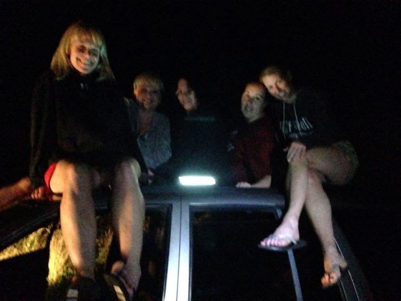Five women sit atop a car smiling in the middle of the night. It is dark outside, but a few small lights illuminate the group.